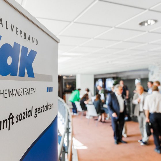 Auf einem Roll-up steht das Logo des VdK NRW mit dem Schriftzug "Zukunft sozial gestalten". Im Hintergrund unterhalten sich mehrere Menschen.