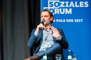 Referent William Wofgramm sitzt auf der Bühne und spricht ins Mikrofon