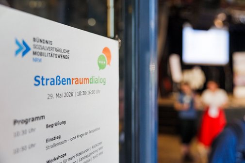 Plakat mit der Aufschrift Straßenraumdialog, verschwommen im Hintergrund der Blick in einen Saal mit Bühne und Menschen.