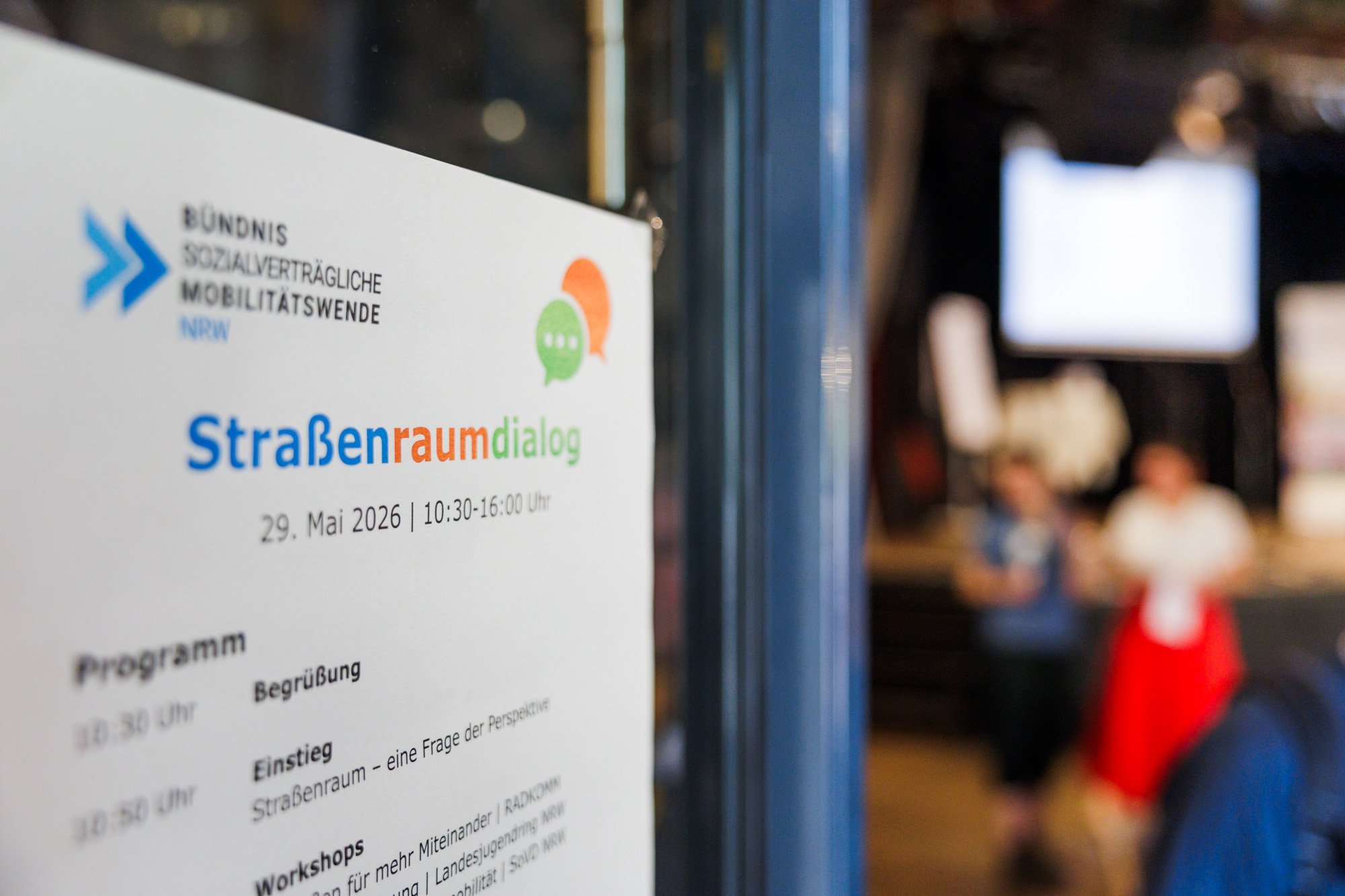 Plakat mit der Aufschrift Stra&szlig;enraumdialog, verschwommen im Hintergrund der Blick in einen Saal mit B&uuml;hne und Menschen. 