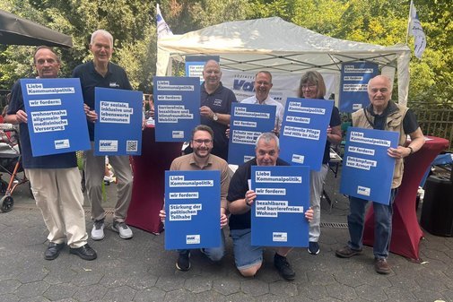 Eine Gruppe Menschen steht und hockt. Alle halten blaue Plakate in den Händen, auf denen in weiß die Forderungen des VdK an die Kommunalpolitik stehen: mehr Barrierefreiheit, bezahlbares Wohnen, gute medizinische Versorgung.