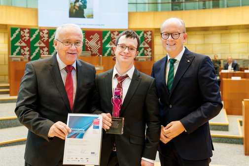 Preisträger Tobias Koddebusch steht mit vilmA-Preis in der Hand zwischen zwei Männern im Anzug. 