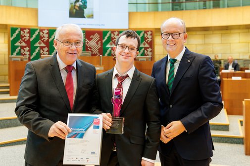 Preistr&auml;ger Tobias Koddebusch steht mit vilmA-Preis in der Hand zwischen zwei M&auml;nnern im Anzug. 