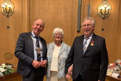 Zwei ältere Herren in dunklen Anzügen stehen rechts und links von einer weißhaarigen Frau in heller Hose und Bluse in einem holzvertäfelten Raum mit hellem Parkett.
