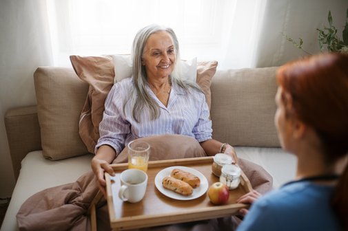 eine Pflegebedürftige bekommt von einer Angehörigen ein Tablett mit einem Frühstück