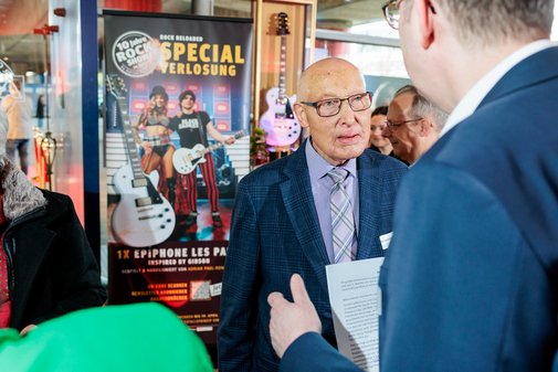 Zwei M&auml;nner mit Sakko unterhalten sich. Im Hintergrund h&auml;ngt ein Plakat mit der Aufschrift "Special Verlosung" und einer E-Gitarre.