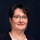 Porträt einer Frau mit kurzen dunklen Haaren und Brille in einem schwarzen Blazer mit weißer Bluse und goldener Halskette vor einem dunkelblauen Hintergrund.