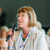Eine ältere Frau mit Brille, kinnlangen hellbraunen Haaren und beigem Blazer spricht in ein Mikrofon, im Hintergrund am Rande zu erkennen: ein Saal voller Menschen.