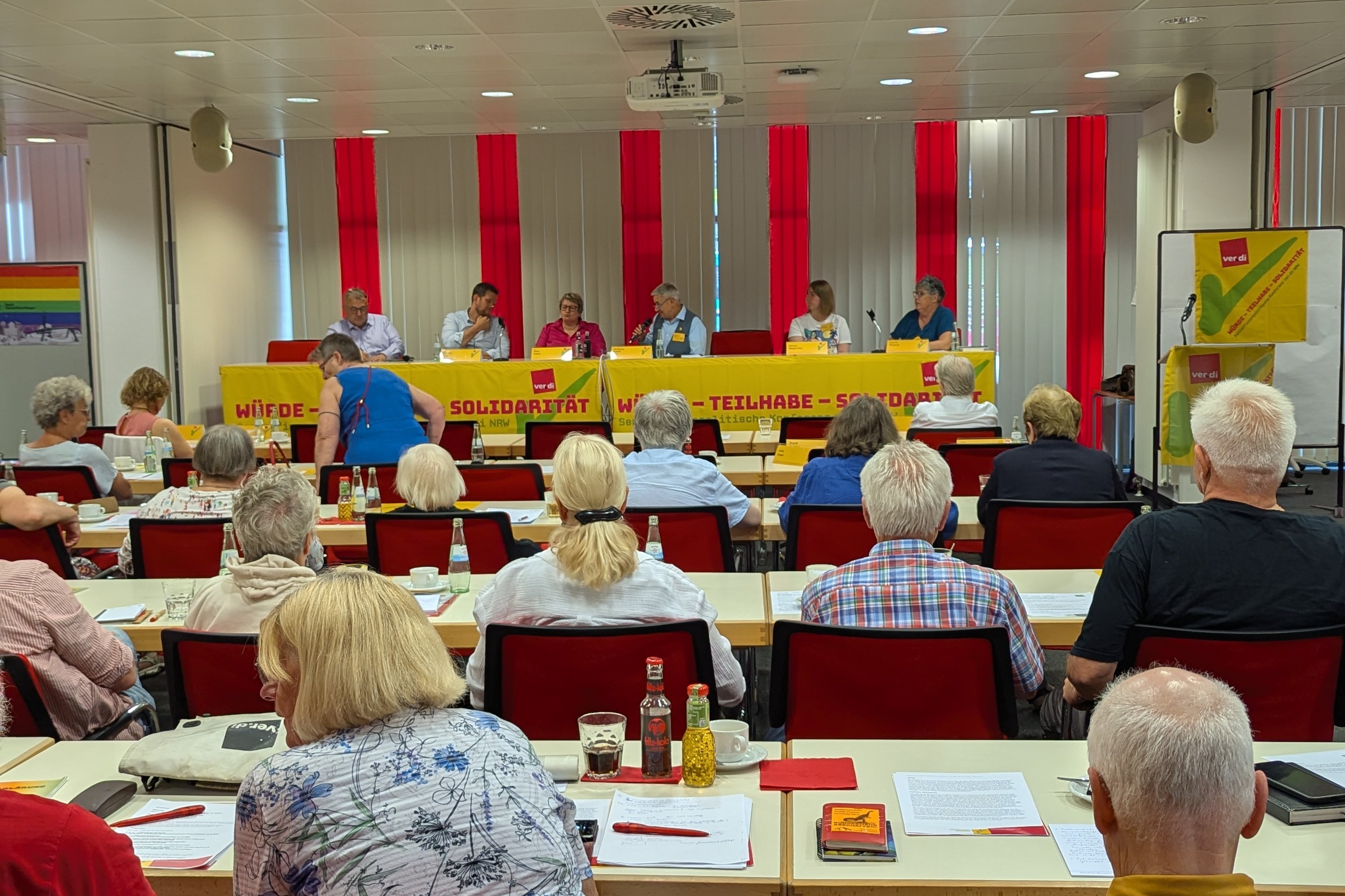 Blick in einen Saal voller Menschen, am Ende eine Bühne mit sechs Männern und Frauen die gemeinsam zur Podiumsdiskussion an einem Tisch mit ver.di-Banner sitzen.