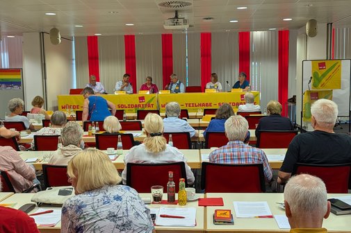Blick in einen Saal voller Menschen, am Ende eine Bühne mit sechs Männern und Frauen die gemeinsam zur Podiumsdiskussion an einem Tisch mit ver.di-Banner sitzen.