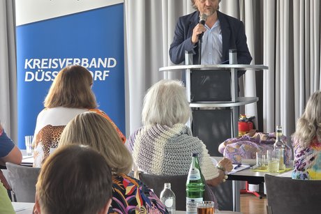 Ein Mann spricht auf einer Bühne in ein Mirkofon, neben ihm ein Roll-Up mit VdK-Logo und Schrift Kreisverband-Düsseldorf.