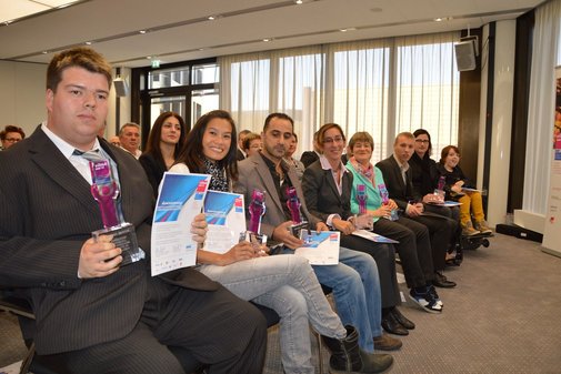 VilmA-Preisträger*innen sitzen in der ersten Reihe eines Saals mit Urkunden und Pokalen auf dem Schoß.