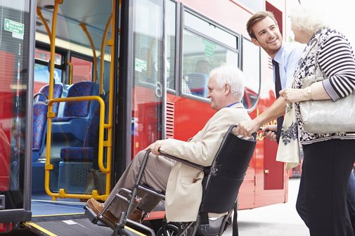 Ein Busfahrer hilft einer &auml;lteren Frau dabei, einen Mann im Rollstuhl &uuml;ber die Rampe in den Bus zu schieben