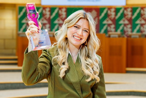 Eine Frau mit langen blonden Haaren h&auml;lt l&auml;chelnd den vilmA-Preis in die H&ouml;he. 