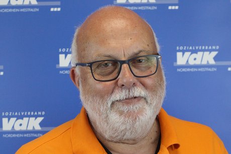 Porträt eines älteren Herren mit dunkelumrandeter Brille, Glatze und kurzem weißen Bart vor einer blauen Wand mit kleinen weißen VdK-Logos.