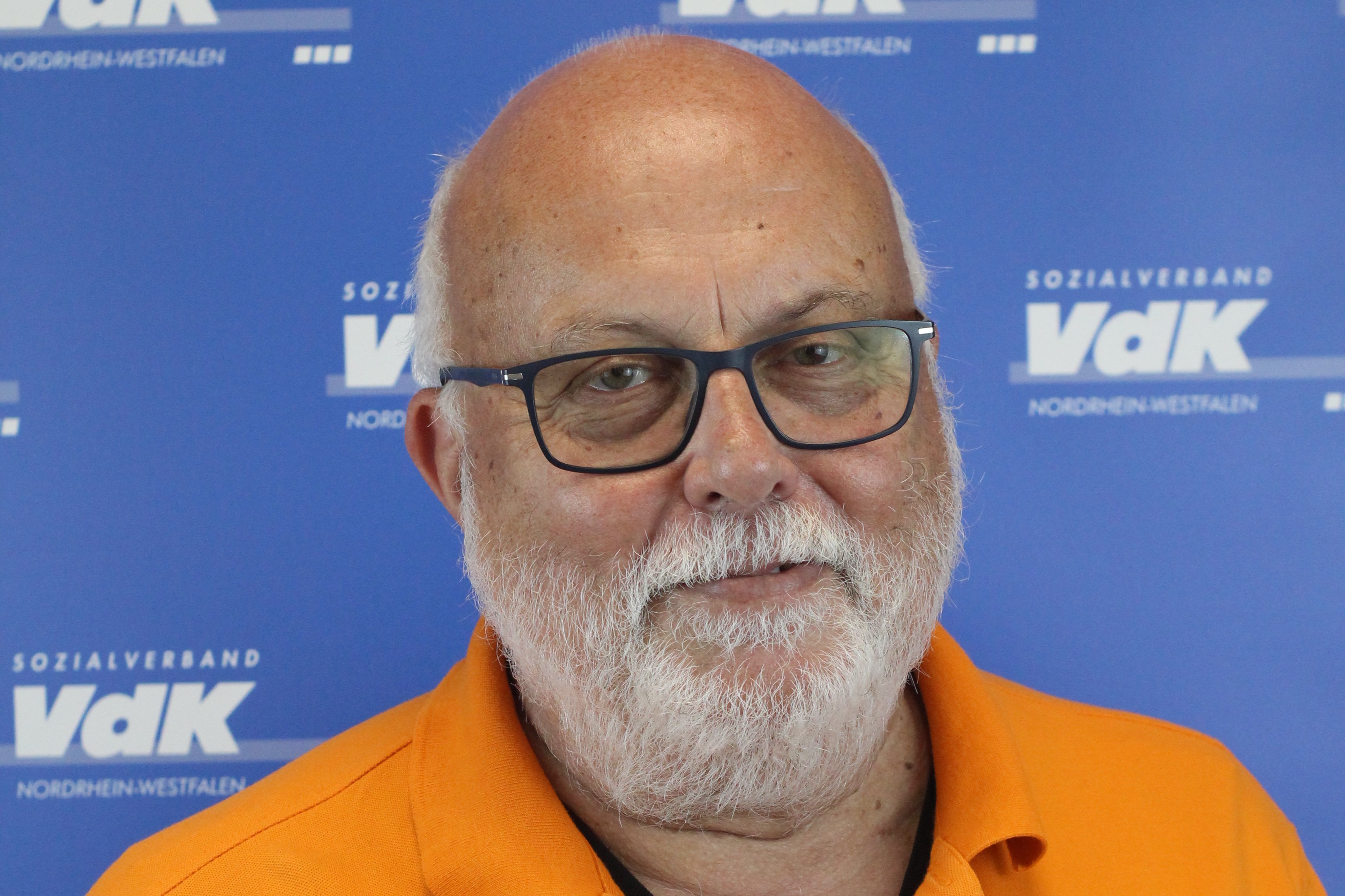 Norbert Zielonka Porträt eines älteren Herren mit dunkelumrandeter Brille, Glatze und kurzem weißen Bart vor einer blauen Wand mit kleinen weißen VdK-Logos.