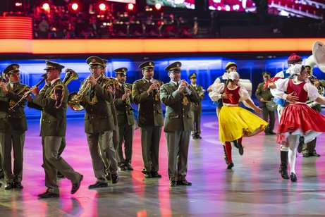 In einer Arena stehen uniformierte Musiker mit Blasinstrumenten. Daneben tanzen T&auml;nzerinnen und T&auml;nzer in Trachten.