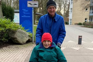 Rolf Buddensieck schiebt seine Frau in einem Rollstuhl eine Straße entlang.