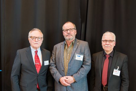 Drei Männer in Anzügen stehen nebeneinander vor einem schwarzen Vorhang.