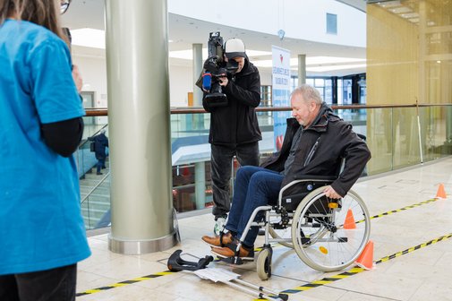 Ein mann im Rollstuhl versucht einen Parcours mit Barrieren abzufahren. Im Hintergrund filmt ein Mann mit Kamera die Aktion.