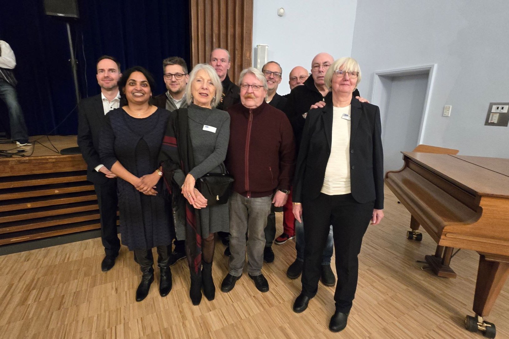 Eine Gruppe von Menschen posiert für ein Foto in einem Saal mit Bühne und Flügel. Im Hintergrund spielt ein Musiker Gitarre.
