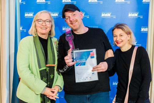 Ein vilmA-Preisträger steht mit Urkunde und Pokal vor einer blauen Wand mit VdK-Logos. Neben ihn Laudatorin und Begleiterin. ner