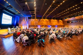 Viele Menschen sitzen in Stuhlreihen in einem großen Saal und schauen Richtung Bühne.