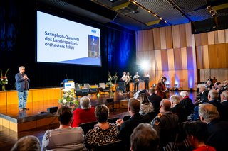 Menschen sitzen im Saal und hören vier Musikern mit Saxophon und Klarinette zu, die rechts auf einer großen Bühne stehen und spielen. Links auf der Bühne steht einer der beiden Gebärdendolmetscher und wartet.