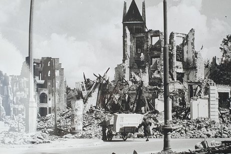 Menschen mit einem Handkarren vor den Tr&uuml;mmern einer zerbombten H&auml;userzeile