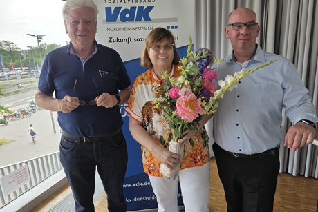 Eine Frau steht mit Blumenstrauß im Arm zwischen zwei Männern vor einem Roll-Up mit VdK-Logo.