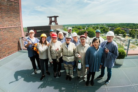 Ein Chor aus Männern und Frauen in typischer Bergmannskluft mit Helmen und Grubenlampen steht auf einem Dach. Im Hintergrund die Kulisse der Zeche mit Förderturm.