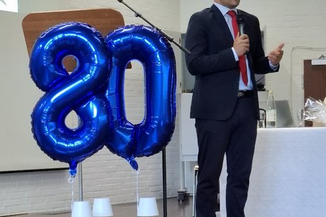 Mahmut Özdemir steht neben einer blauen Luftballon-80 am Rednerpult auf einer Bühne.