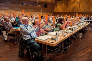 Delegierte sitzen an langen Tischen in einem Saal und halten Stimmzettel in die Luft.