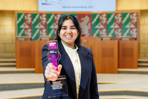 Eine junge Frau mit langen dunklen Haaren in blauem Sakko und heller Bluse streckt den vilmA-Preis Richtung Kamera.