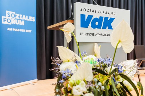 Blick auf die B&uuml;hne mit blumengeschm&uuml;cktem Rednerpult und VdK-Logo und einem Roll-Up mit Schriftzug "Soziales Forum" im Hintergrund