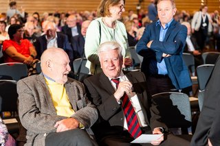 Zwei Männer sitzen im bestuhlten Saal der Stadthalle und unterhalten sich. Hinter ihnen im Bild stehen und sitzen weitere Gäste.