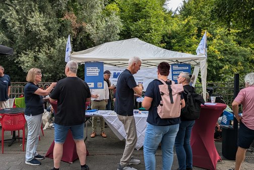 Einige Menschen haben sich rund um den weißen Pavillon des VdK versammelt. Im Hintergrund sieht man in weißer Schrift auf blauem Grund Plakate mit Forderungen an die Kommunalpolitik.