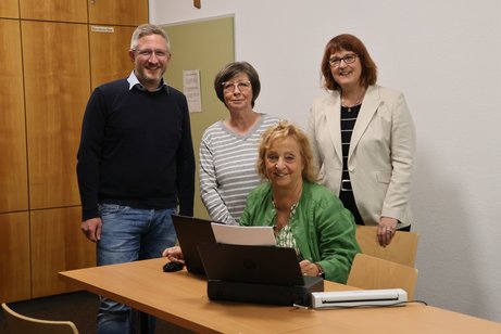 Vier Personen stehen und sitzen gemeinsam in einem Raum mit Holzwänden und weißen Wänden. Eine Frau sitzt an einem Tisch mit einem Laptop und einem kleinen Scanner, drei weitere Personen stehen hinter ihr. Alle blicken freundlich in die Kamera. 