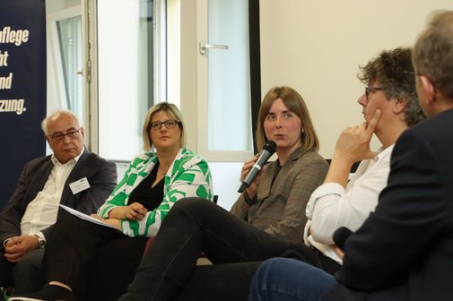 Fünf Menschen sitzen bei einer Podiumsdiskussion nebeneinander. Die Frau in der Bildmitte spricht gerade in ein Mikrofon. Die anderen hören ihr mit ernsten Mienen zu. 