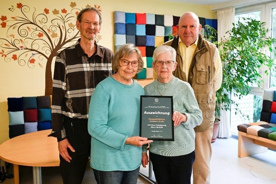 Zwei M&auml;nner und zwei Frauen stehen als Gruppe in einem Raum beisammen. Die beiden Frauen halten gemeinsam eine gerahmte Urkunde in der Hand. 