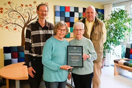 Zwei Männer und zwei Frauen stehen als Gruppe in einem Raum beisammen. Die beiden Frauen halten gemeinsam eine gerahmte Urkunde in der Hand.