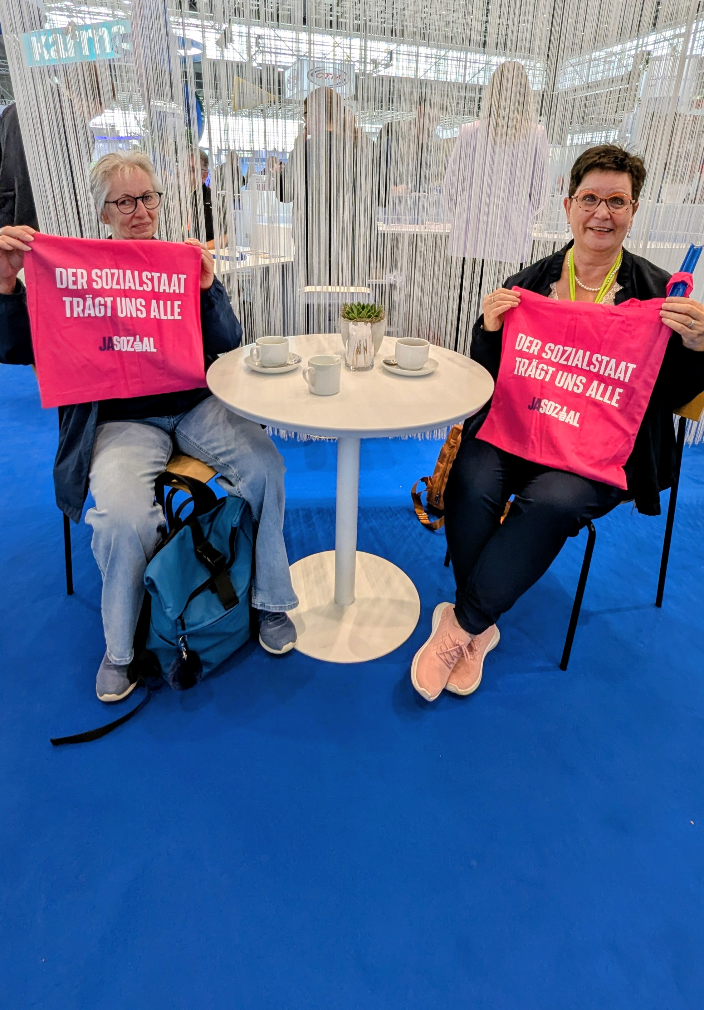 Zwei Frauen sitzen an einem Tisch des VdK-Messestands und halten pinke Einkaufstaschen hoch mit Slogan: Der Sozialstaat trägt uns alle. 