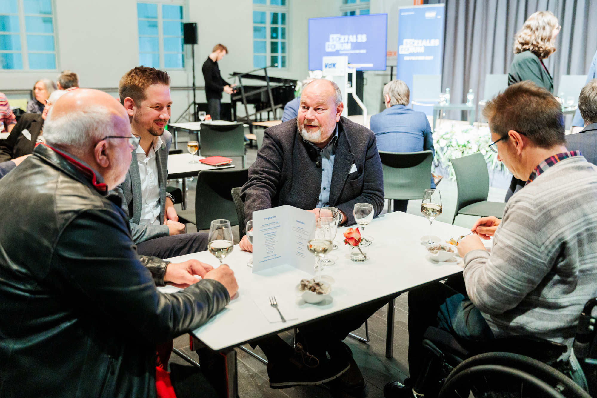 Gäste sitzen gemeinsam an einem Tisch im Gespräch. Rechts im Rollstuhl SoVD-Abteilungsleiter Dr. Michael Spörke.