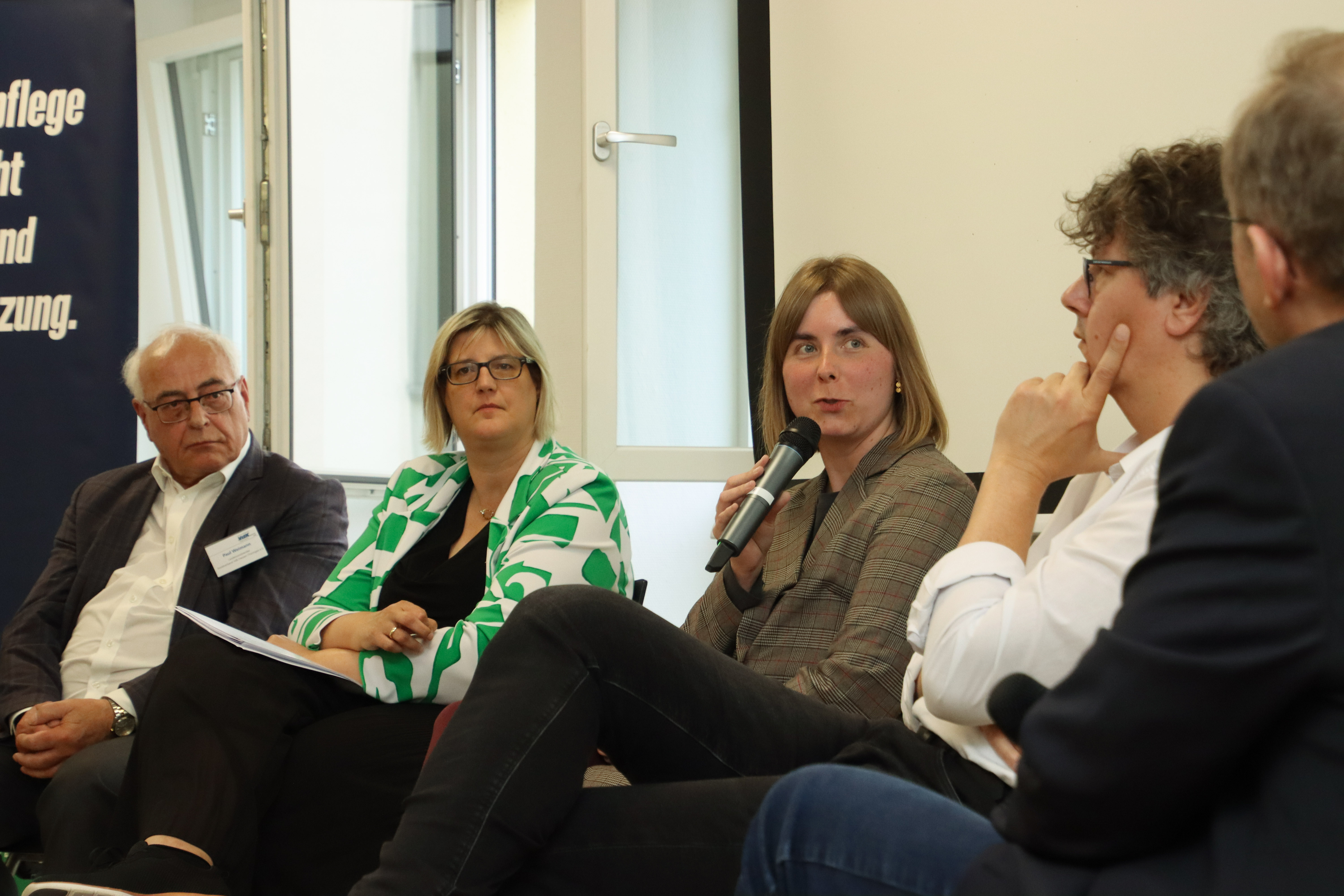 Fünf Menschen sitzen bei einer Podiumsdiskussion nebeneinander. Die Frau in der Bildmitte spricht gerade in ein Mikrofon. Die anderen hören ihr mit ernsten Mienen zu.