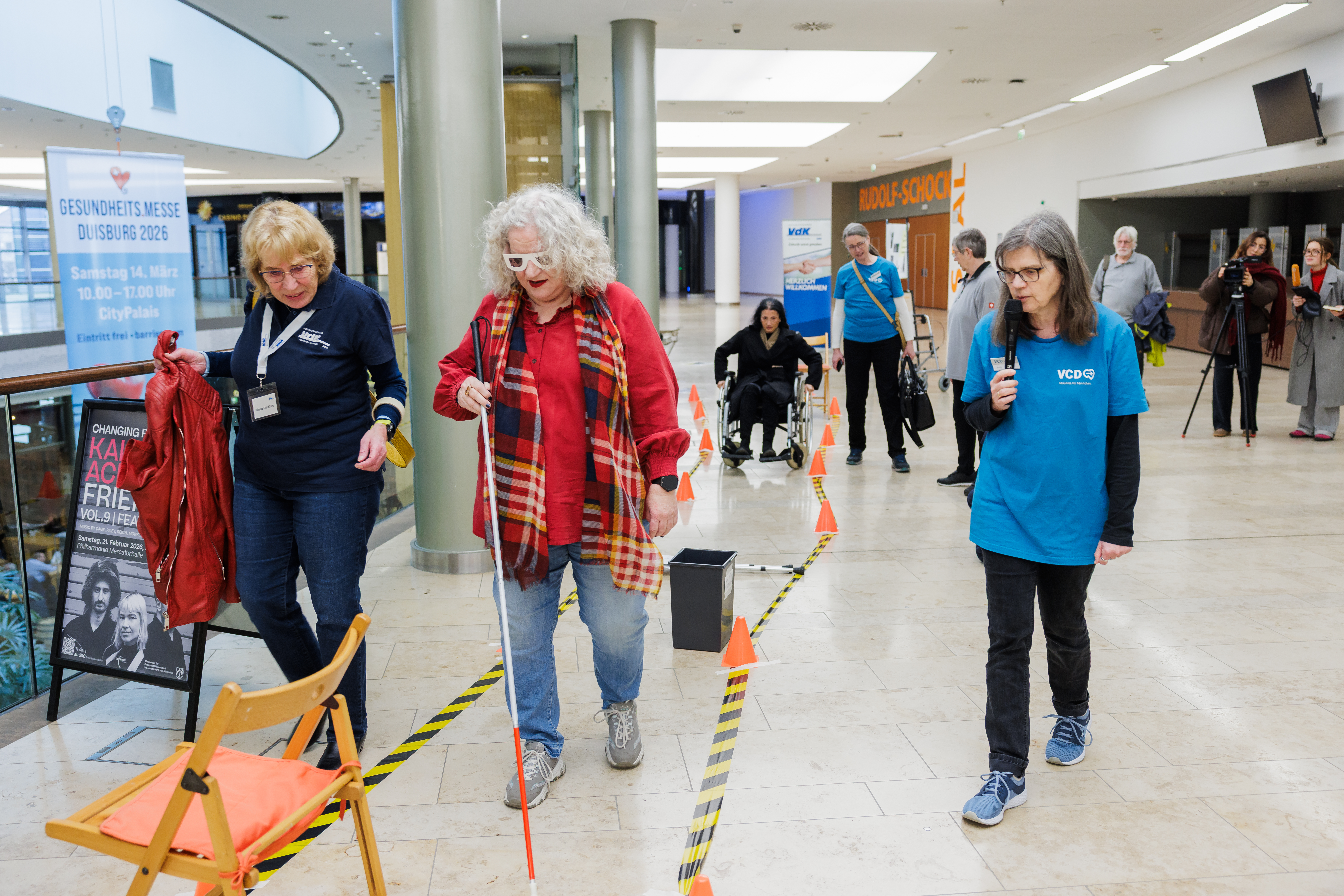 Eine Frau mit dunkelget&ouml;nter Brille versucht mit Blindenleitstock einen Hindernisparcours abzulaufen. 