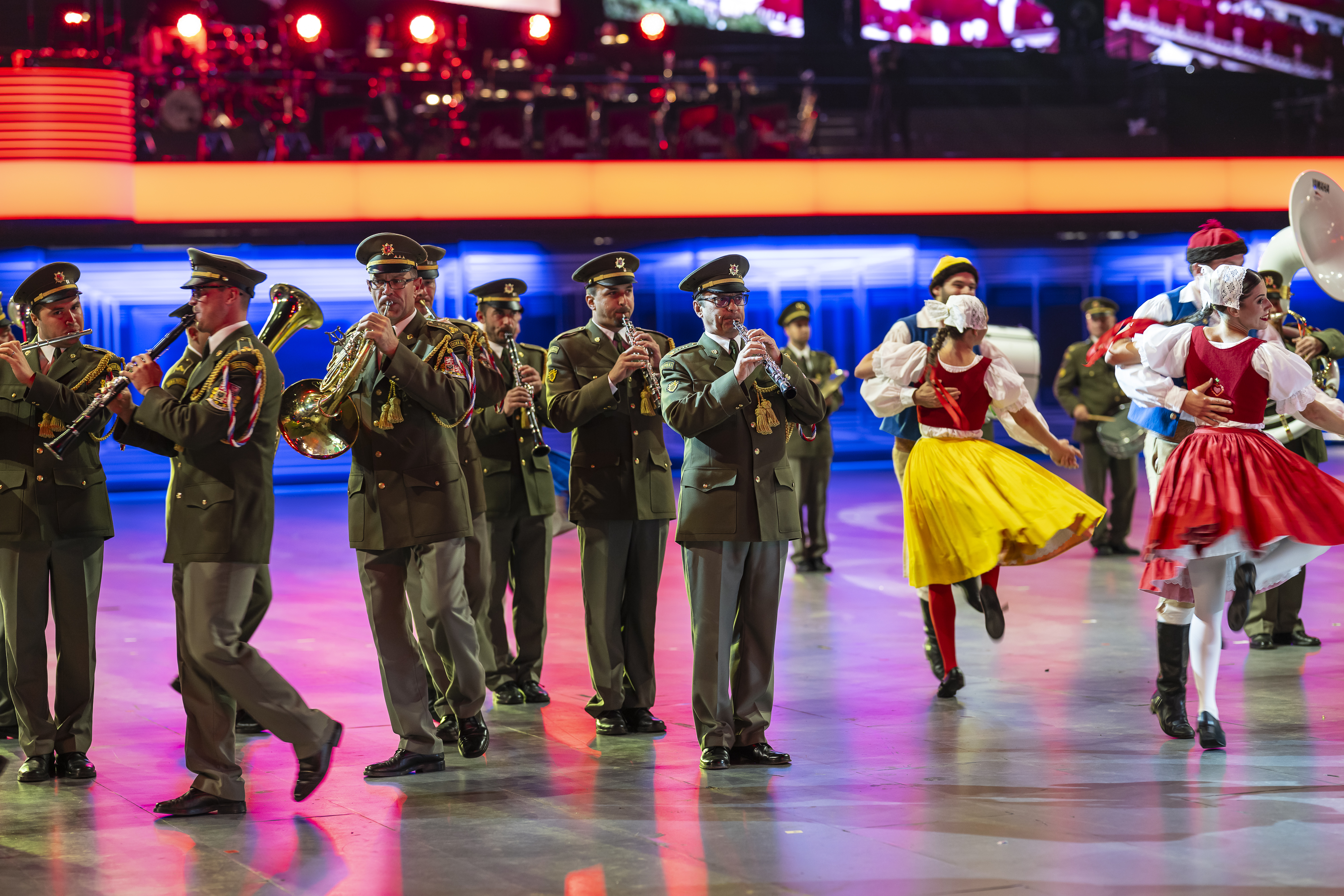 In einer Arena stehen uniformierte Musiker mit Blasinstrumenten. Daneben tanzen Tänzerinnen und Tänzer in Trachten.