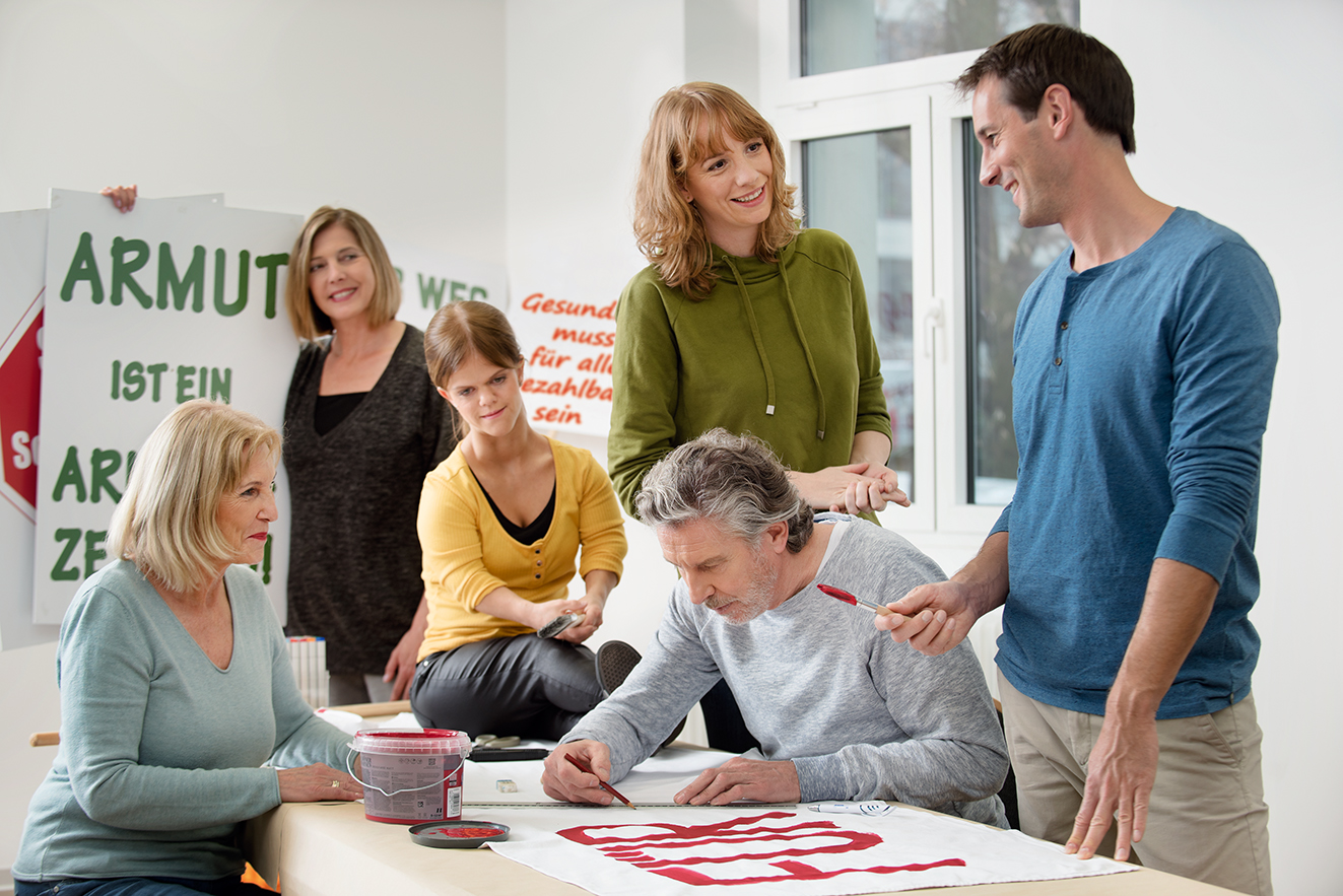 Eine Gruppe unterschiedlicher Menschen - Männer, Frauen, Alte und Junge, mit und ohne Behinderung, malt gemeinsam Demo-Plakate.