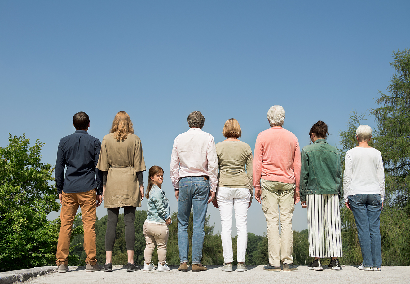 Menschen verschiedenen Alters mit und ohne Behinderung stehen nebeneinander, wenden dem Betrachter den R&uuml;cken zu. Eine kleinw&uuml;chsige junge Frau dreht sich zum Betrachter um.