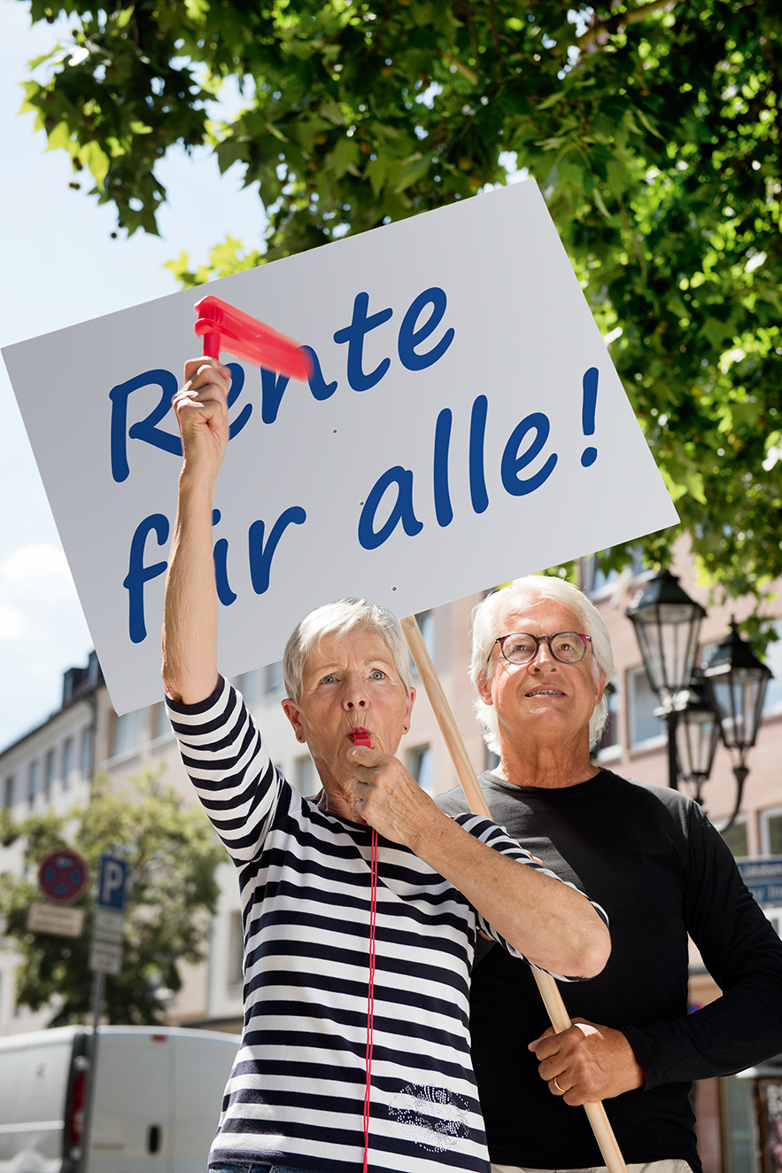 Seniorenpaar auf einer Renten-Demo Eine ältere Frau und ein älterer Mann mit einer Demopappe mit der Aufschrift "Rente für alle!". Die Frau bläst in eine Trillerpfeife, hält eine Ratsche in der Hand.