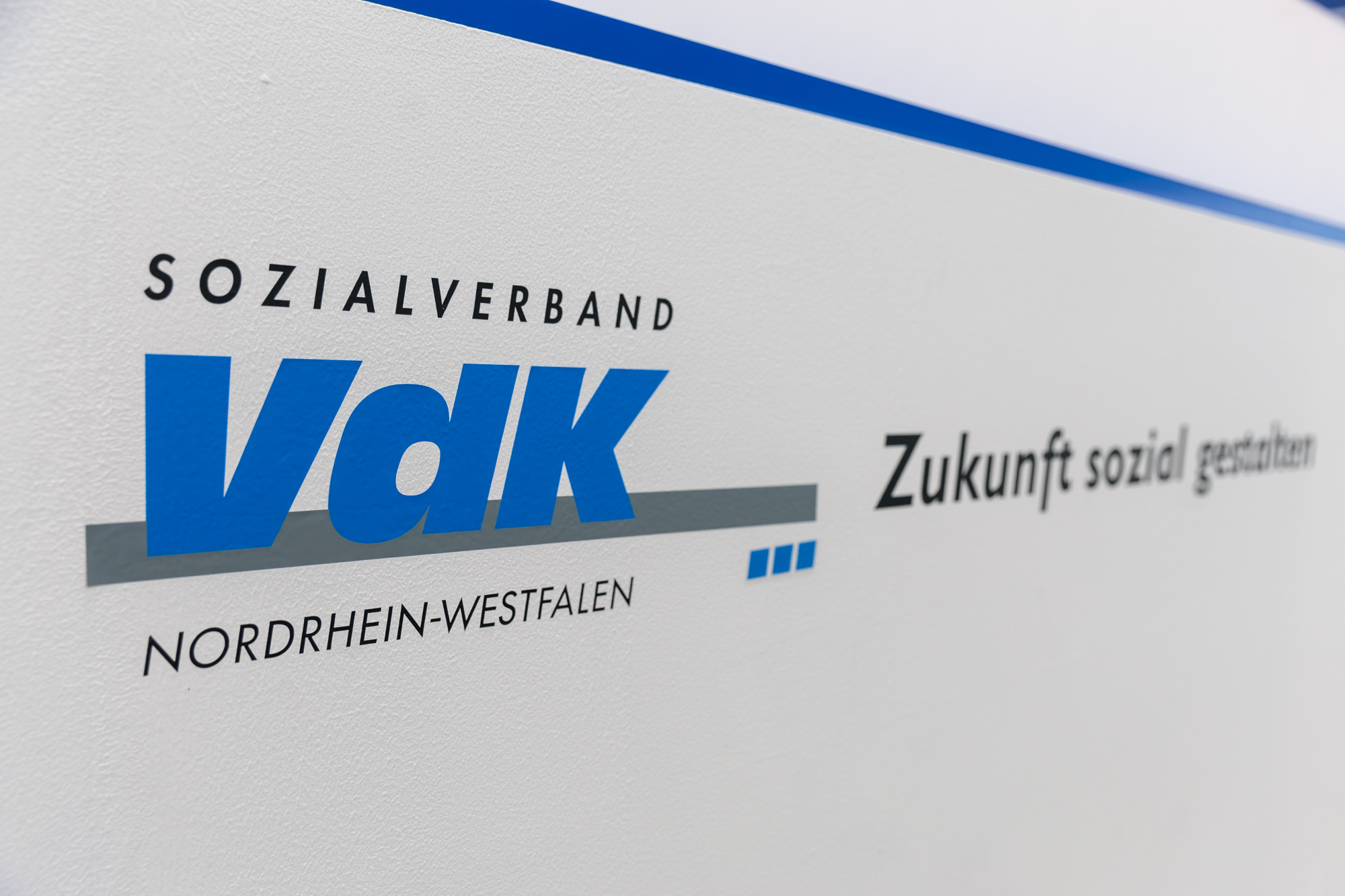 Weiße Wand mit blauem Logo des VdK-NRW und Slogan: Zukunft sozial gestalten